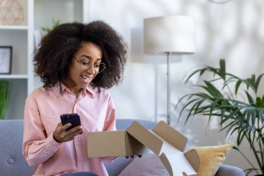 Kanepede akıllı telefon kullanırken gülümseyen kadın paketi açıyor. Bitkiler ve dekorlu ev ortamı.