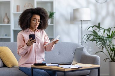 Modern oturma odasında kişisel hesapları yöneten akıllı telefon ve laptop kullanan bir kadın makbuzu kontrol ediyor.