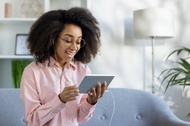 Evde kulaklık takan ve gülümseyen bir kadın kanepeye oturmuş online içeriğin tadını çıkarıyor.