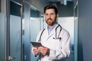 Beyaz önlüklü steteskop takmış, elinde tablet olan bir erkek doktor. Modern hastane koridorunda kendine güvenen bir profesyonel, gülümsüyor. Sağlık, tıbbi uzmanlık, teknoloji entegrasyonu.