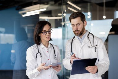 Beyaz önlüklü iki doktor, çağdaş tıp ofisinde hasta verilerini bir pano ve tablet üzerinde gözden geçiriyorlar. İş toplantısında işbirliği yapan odaklı sağlık uzmanları