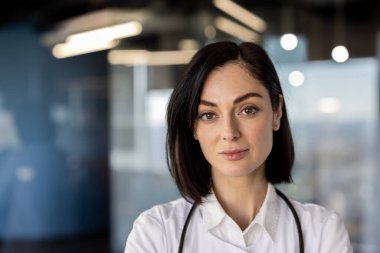 Modern tıp ofisinde steteskopla dikilen kendine güvenen kadın doktor portresi. Çağdaş klinik ortamında ciddi ifadesi olan profesyonel sağlık çalışanı.