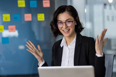 Modern bir ofiste kendine güvenen bir iş kadını çevrimiçi bir toplantı sırasında heyecanını dile getiriyor. Gözlüklü ve resmi takım elbiseli bir kadın, dizüstü bilgisayar kullanırken coşkuyla el kol hareketi yapıyor..