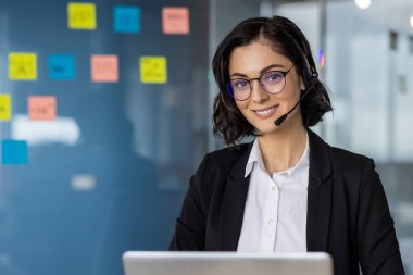 Modern ofis ortamında çalışan, kulaklık takan gülümseyen bir kadın. Renkli yapışkan notlarla çevrili müşteri desteği sağlayan profesyonel çağrı merkezi temsilcisi.