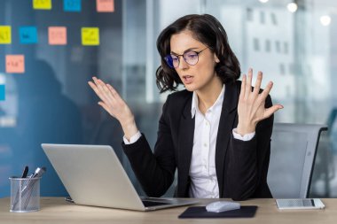 Modern ofis ortamında dizüstü bilgisayar üzerinde çalışırken hayal kırıklığını ifade eden iş elbiseli kızgın kadın. Not kağıtları arka plandaki cam duvarda görünüyor..