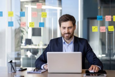 Ofisin içinde dizüstü bilgisayarla çalışan başarılı gülümseyen adam, klavyede yazan iş adamı sonuçlarından memnun, iş yerinde takım elbiseli olgun bir patron..
