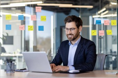Ofisin içinde dizüstü bilgisayarla çalışan başarılı gülümseyen adam, klavyede yazan iş adamı sonuçlarından memnun, iş yerinde takım elbiseli olgun bir patron..