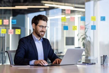 Ofisin içinde dizüstü bilgisayarla çalışan başarılı gülümseyen adam, klavyede yazan iş adamı sonuçlarından memnun, iş yerinde takım elbiseli olgun bir patron..