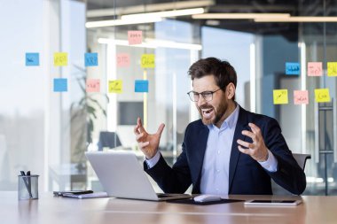 İş elbisesi giymiş kızgın bir adam ofisin içinde dizüstü bilgisayara bağırıyor, iş yerindeki olgun bir işadamı şirket performansından memnun değil, online çalışanlarla tartışıyor..