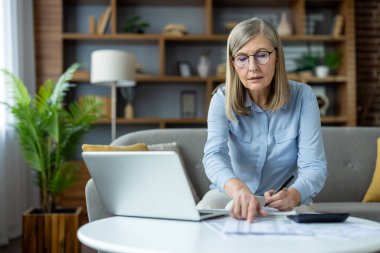 Evde dizüstü bilgisayarda çalışan olgun bir kadın masadaki belgelere işaret ediyor. Evrak işlerine odaklandım, oturma odasındaki koltukta oturdum. Konforlu ev ortamında uzaktan çalışma.