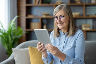 Evde dijital tablet kullanan, kanepede oturan gülümseyen son sınıf öğrencisi kadın. Teknolojinin tadını çıkarmak, aile ve arkadaşlarla bağlantıda kalmak. Doğal ışık, kitaplık ve yeşil bitkilerle modern oturma odası..