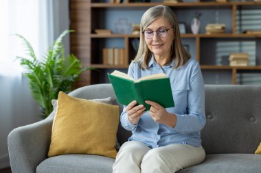 Yaşlı bir kadın rahat bir kanepede kitap okuyup kitap okumaktan zevk alıyor. Kitap rafları ve bitki süslemeleriyle dolu, rahat bir oturma odasında.