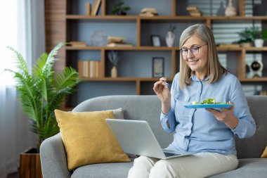 Evde dizüstü bilgisayarla çalışırken kıdemli kadın salata yiyor. Modern iç dekorasyonlu rahat bir ofis. Emeklilik, uzak çalışma ve sağlıklı yaşam kavramları.