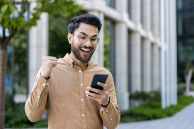 Hispanik adam şehir dışında akıllı telefon kullanırken heyecanlanıyor ve başarıyı kutluyor. Gözlüklü, gülümseyen ve yumruğunu zaferle gösteren sıradan bir adam..