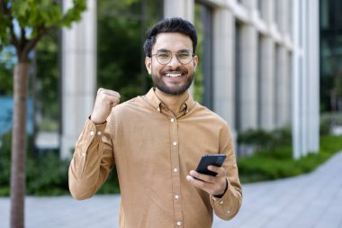 Şehir ortamında akıllı telefon tutan, gözlüklü, kendine güvenen İspanyol bir adam, başarıyı yumruk pompasıyla kutluyor. İş ortamında profesyonel ve olumlu duygular.