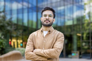 Modern cam binanın dışında kolları çapraz duran gözlüklü, kendine güvenen İspanyol bir adam kameraya bakıyor. Kavram işi, başarı, güven