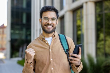 Akıllı telefonu elinde tutan İspanyol öğrenci mutlu görünüyor ve kampüs binasının dışında başarıyı kutluyor. Sırt çantalı genç adam heyecan gösteriyor. Başarı, eğitim ve öğrenim hayatı kavramı.
