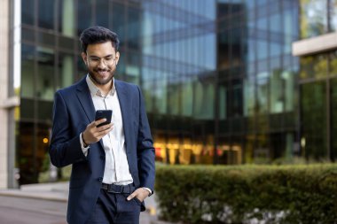 Modern ofis binasının dışında akıllı telefon kullanan resmi giyinmiş İspanyol işadamı. Gülümseyen, teknolojiyle uğraşan ve kendine güvenen bir adam. Arkaplanda kurumsal çevre cam cephesi.