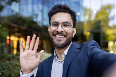 İspanyol işadamı gözlük takıyor, takım elbise giyiyor ve modern ofis binasının önünde kameraya gülümsüyor ve el sallıyor. İletişim kavramı, iş dünyası profesyoneli ve şehir yaşam tarzı.