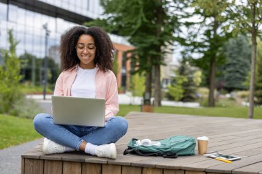 Genç kız öğrenci kampüste parkta bankta oturuyor, dizüstü bilgisayar kullanıyor, gülümsüyor, günlük giysiler giyiyor, temiz havanın tadını çıkarıyor, internetten ders çalışıyor.