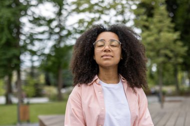 Genç bayan öğrenci, sakin bir park ortamında meditasyon yapmak için ara veriyor, gözleri kapalı ve rahat bir ifadeyle. Manzara huzurlu ve huzurlu bir atmosferi yakalar..