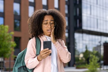 Genç kız öğrenci endişeli görünüyor. Akıllı telefonu kontrol ediyor. Görüntü, endişeyi, dış mekanı ve arka plandaki modern kampüs binalarını yakalıyor. Endişe, şaşkınlık ve belirsizliği gösterir..