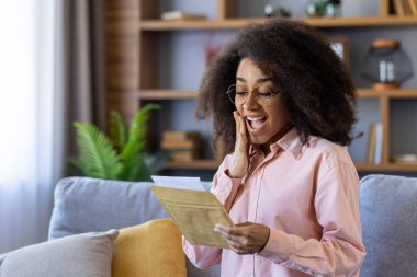 Gözlüklü, heyecanlı bir kadın evde mektup okuyor. Sürpriz ve neşe dolu bir ifadeyle. Zarf tutarken gerçek mutluluğu ve olumlu duyguları gösterir.