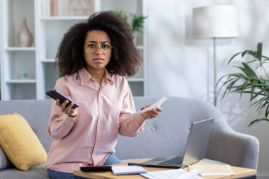 Kanepede oturmuş, elinde telefon ve kağıtlarla kendini stresli hisseden genç bir kadın. Endişeli ifade maddi ya da işle ilgili sorunları akla getiriyor. Laptop ve belgeleri sehpanın üzerine kurun.