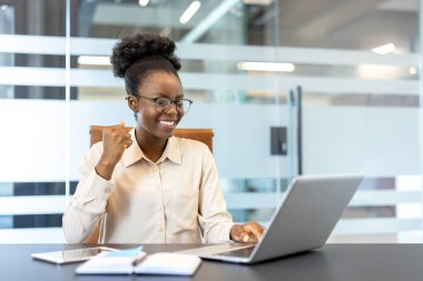 Modern ofiste dizüstü bilgisayarda çalışan mutlu profesyonel kadın. İş kadını gözlük takıyor, özgüvenle gülümsüyor ve başarıyla yumruk tokuşturuyor. Doğal ışıklandırma ve cam duvarlı ofis alanı.