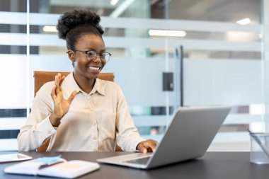 Gülümseyen iş kadını laptopunda video görüşmesi yapıyor. Profesyonel bir kadın internette el sallıyor. Uzaktan çalışma, teknoloji, iletişim ve iş bağlantıları kavramı.