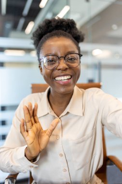 Modern ofis ortamında gülümseyen ve el sallayan arkadaş canlısı bir kadın. Profesyonel iletişim sanal toplantı yoluyla sağlanır. Olumlu, uzak iletişim için teknolojiyi kullanarak etkileşim kurma.