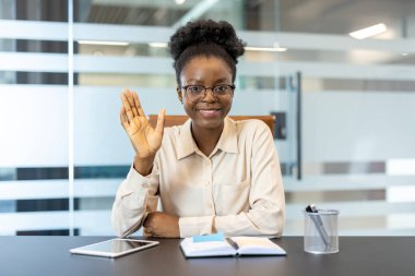 Modern ofis ortamında bir video görüşmesi sırasında iş kadını el sallıyor. Not defteri, tablet ve kalem tutacağı masanın üstünde. Profesyonel ve dostane çalışma ortamı.