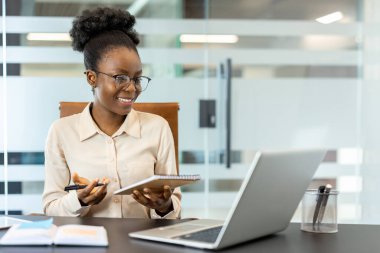 Gülümseyen iş kadını modern ofiste elinde defter ve kalemle verimli bir video görüşmesi yaptı. Sanal toplantı ya da çevrimiçi konferans için dizüstü bilgisayarla iletişim kuran kendinden emin profesyonel