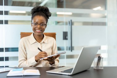 Kendine güvenen genç iş kadını, modern ofiste dizüstü bilgisayarla masa başında not alıyor. Gülümseyen kadın profesyonel planlama ve organize çalışma. Üretkenlik, başarı ve kariyer büyüme kavramı.