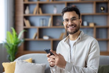 Modern oturma odasında akıllı telefon kullanan gülümseyen genç adam rahat bir kanepeye oturdu. Parlak ve rahat ofis dekorasyonu, ahşap kitaplık ve yeşil bitki arka planı.