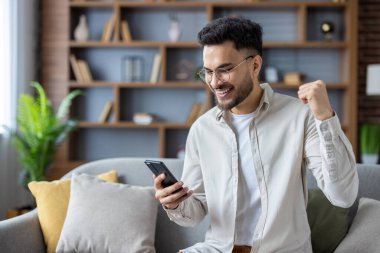 Adam akıllı telefona bakarken koltukta oturmuş heyecanını dile getiriyor. Arka planda ahşap raflar ve bitkiler olan modern bir ev. Başarı, mutluluk ve başarı kavramı.