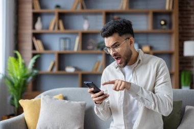 Modern oturma odasında akıllı telefon kullanırken hayal kırıklığını ifade eden bir adam. Kavram, reaksiyon, teknoloji, duygu ve evle ilgilidir. Arka plandaki raflar ve bitkiler rahat ortamı güçlendirir.