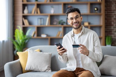 Evdeki genç Müslüman adamın portresi, kanepede oturuyor, elinde bir kredi kartı ve cep telefonu, kameraya gülümsüyor..