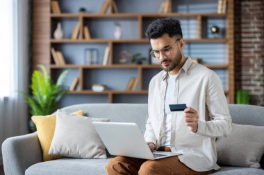 Kanepede oturan adam dizüstü bilgisayar kullanıyor ve modern oturma odasında rafları ve bitkileri olan online alışveriş için kredi kartı tutuyor.