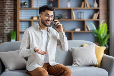 Genç adam kanepede oturup telefonla konuşuyor, gülümsüyor ve el kol hareketi yapıyor. Kitaplığı ve arka planda bitkileri olan modern oturma odası. Rahat ve rahat bir ortam..