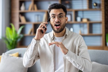 Evde cep telefonuyla konuşan sinirli bir adam üzgün ve sinirli bir ifade sergiliyor. Duygu, iletişim, çatışma, kapalı alan, ev düzeni