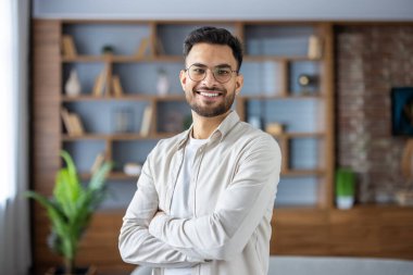 Genç Hintli bir adamın portresi, gözlüklü ve günlük kıyafetlerle, evde, kollarını göğsünde kavuşturmuş kameraya gülümsüyor..