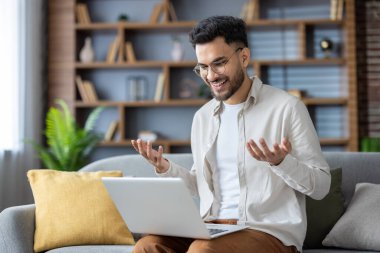 Hintli gülümseyen genç adam evdeki kanepede oturuyor, dizüstü bilgisayarını kucağında tutuyor ve el hareketleriyle video konuşmaları yapıyor..