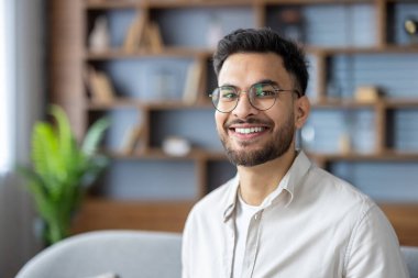 Gözlüklü ve sıradan giyinmiş gülümseyen genç bir profesyonelin portresi. Arkaplanda dekoratif eşyalar ve yeşil bitkilerle modern ofis kitaplığı yer alıyor.