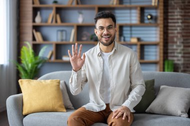 Gözlüklü, gülümseyen ve video görüşmesinde el sallayan mutlu bir adam ev ofisindeki koltukta oturuyor. Modern dekorlu samimi oturma odasında arkadaşça görüntülü sohbet.