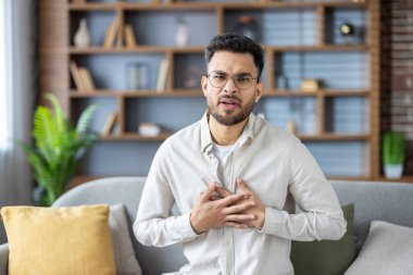 Genç Hintli bir adamın portresi kanepede oturuyor ve elleriyle göğsünü tutuyor, şiddetli bir acı hissediyor, endişeli bir şekilde kameraya bakıyor..