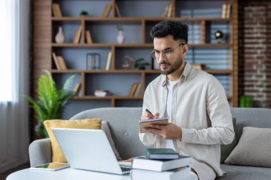 İnternetten öğreniyorum. Hintli genç bir erkek öğrenci elinde bir kitapla kanepede oturur, bir deftere notlar alır, gerçekten uzaktan ders dinler..