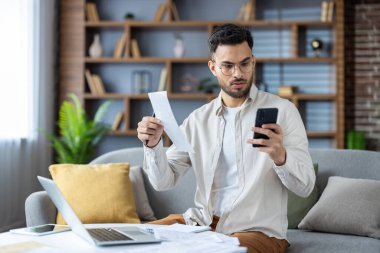 Hintli genç adam evdeki kanepede oturuyor, elinde mektup, makbuz ve endişe içinde cep telefonuna bakıyor, faturaları kontrol ediyor..