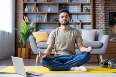 Hintli bir adam oturma odasındaki sarı bir paspasın üzerinde rahatça meditasyon yaparken görülüyor. Çevre, modern mobilyaları ve doğal ışığı olan huzurlu ve sakin bir ev öneriyor..