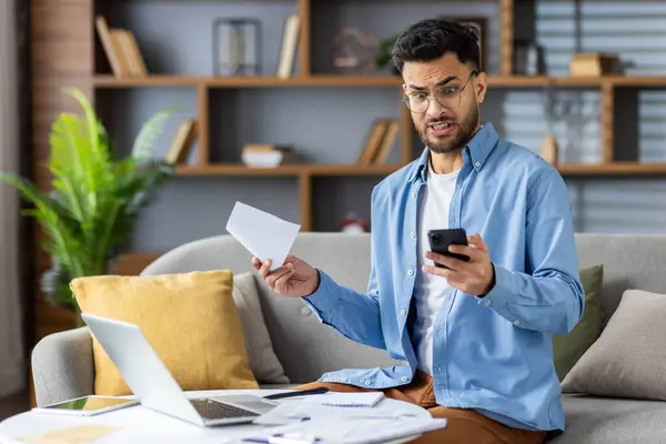 stock image Young man sitting on couch in home office holding phone and financial paperwork looking stressed. Concept of financial problems, stress, home office, remote work